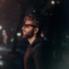 side profil eof man with mahogany colur hair and beard, wearing sunglasses, against a night time background