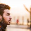 Side profile of man with deep auburn hair colour and beard wearing earphones and rucksack-against a blurred background
