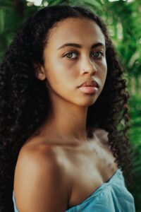 Beautiful young black woman with long wavy hair in a sleeveless dress revealing bare shoulders and beautiful skin, standing againats a natural green backgroundwoman