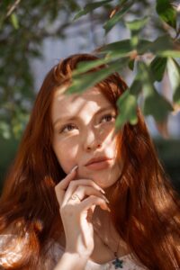 Dark Copper brown hair colour on woman with long, straight, copper colour hair, standing in the shadow of a leafy branch