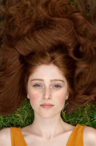 Deep auburn hair dye , image of freckled woman's face, wearing sleeveless orange dress, lying in the grass with deep auburn hair colour splayed all around her face