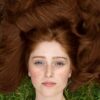 Deep auburn hair dye , image of freckled woman's face, wearing sleeveless orange dress, lying in the grass with deep auburn hair colour splayed all around her face, she's probably hennaed her her with pure henna, the best sensitive scalp hair dye UK