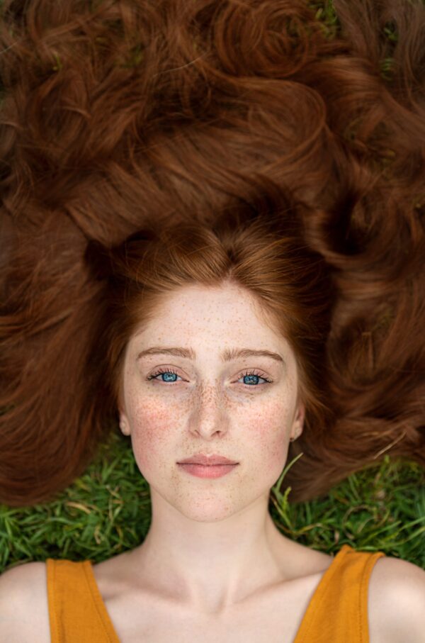 Deep auburn hair dye , image of freckled woman's face, wearing sleeveless orange dress, lying in the grass with deep auburn hair colour splayed all around her face, she's probably hennaed her her with pure henna, the best sensitive scalp hair dye UK