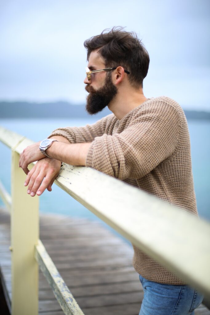 man with dark brown black hair and beard and beige cream jumper standing at a fence and looking out to sea. Hair colour for men dark brown black