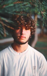 Henna is for guys too - Man with curly hair coloured naturally , wearing a round neck white T shirt, standing in a natural background by a tree branch
