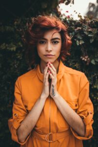 Asian looking woman wearing light orange blouse top with short wavy hair and auburn hair colour, holding her hands together just below her chin