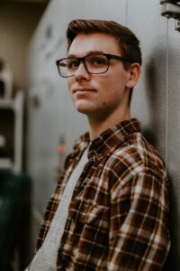 man with light brown hair colour wearing glasses, standing against a wall, wearing a brown and cream check shirt with gray T shirt underneath