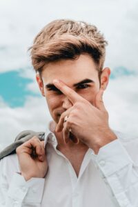 man with naturally coloured light brown hair standing against natural background, wearing white shirt, stringing his jacket across the back of one shoulder, raised hand towards his face and posing - holding one finger diagonally across his face from the corner of his inner eyebrow down across his nose ti