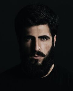 Man with conditioned beard and hair from Mens Natural Beard Oil; bearded dark haired man wearing dark T shirt with a serious gaze