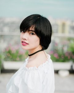 Plant hair dye for short hair will turn hair auburn with henna, and then it will turn your auburn hennaed hair brown through to black hair colour with indigo; woman in white top wearing a black choker, with short black hair.