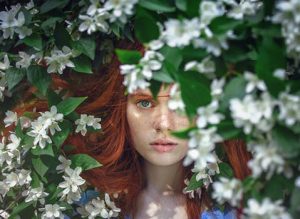 Natural beauty products, plant based, Renaissance Henna; picture of young woman with auburn hair, dace opartially obscured by green leaves and white flowers.