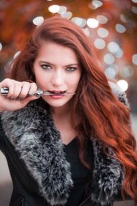 Woman dressed in black coat with grey fur neckline, long auburn hair draping over her shoulder, looking straight at the camera and appearing to hold a utensil like a crockery knife to her mouth. Her hair colour is a stunning natural light reddish brown hair colour which can be attained with Amla & Acacia Catechu natural hair dye 
