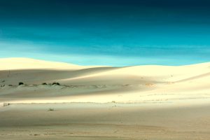 Image of fine white sandy desert gentle rolling landscape against a backdrop turquoise blue sky. The white clay for natural pore tightening often comes from mountains close to these desert regions 