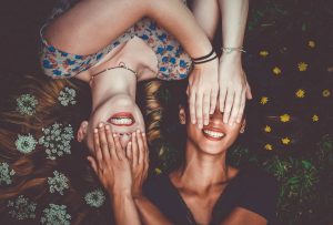 natural hair care, photo of two young women with long brown hair lying on the ground with hair spread out around them, decorated with flowers, covering each others eyes