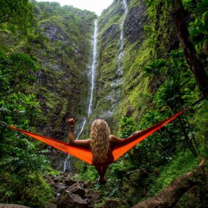 Woman with long hair down her back on red hammock facing a rock face with trickling waterfall