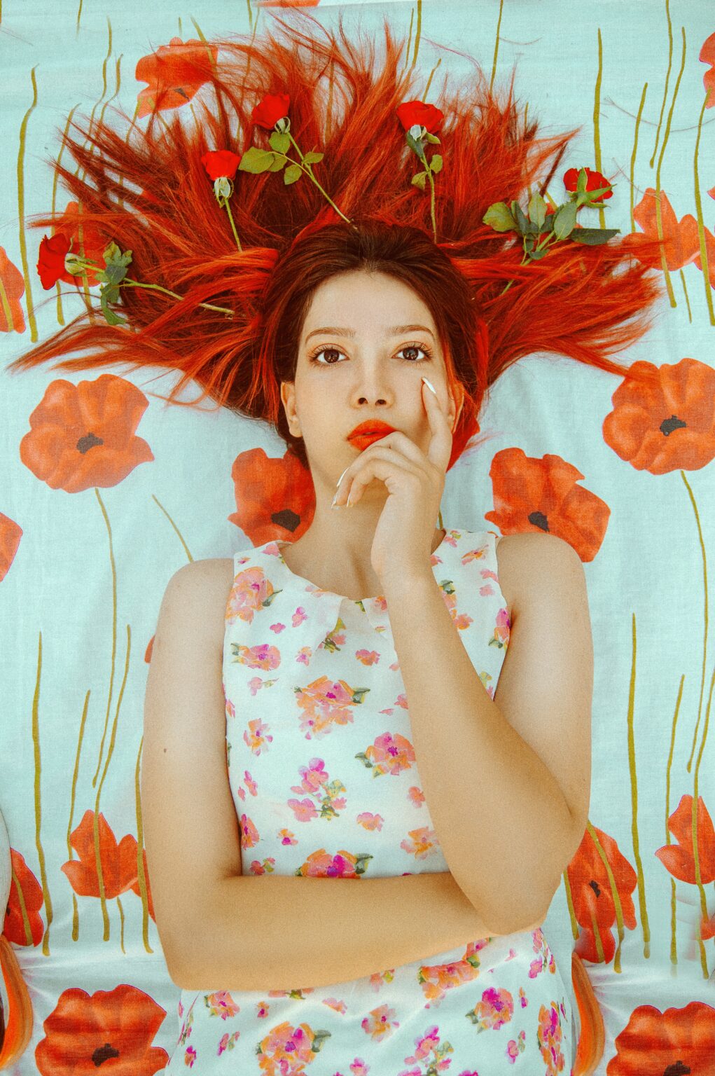 woman with henna hair pondering with hand and finger on her face, against a background of painted wall with red flowers