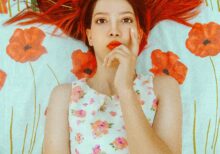 woman with henna hair pondering with hand and finger on her face, against a background of painted wall with red flowers