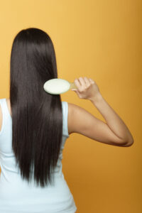 back of woman with long silky healthy hair conditioned by organic hair oils holding a hair brush in her hand and brushing her beautiful hair. This is about looking after your hair with healthy hair oils.