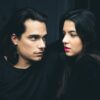 man and woman with black hair, facing each other, against a dark background, both wearing black tops