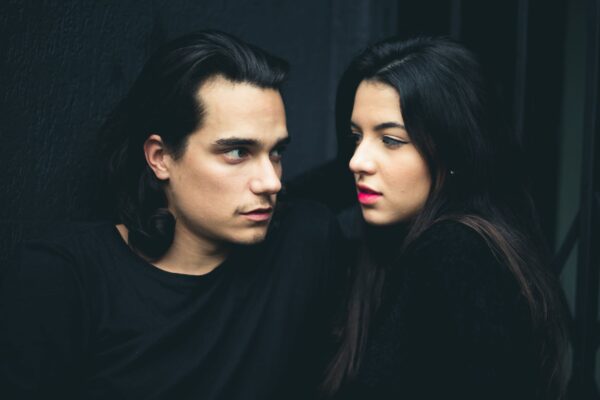 man and woman with black hair, facing each other, against a dark background, both wearing black tops