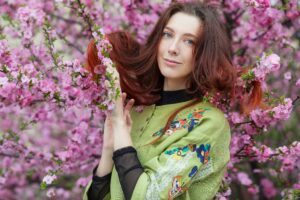 Top half of Woman with shoulder length reddish brown hair wearing a green top over a black turtle neck, standing in front of pink blossoms