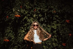 woman with long blonde curls flowing over her shoulders wearing dark sunglasses and sleeves white vest, standing in front of green bush with deep green foliage and red berries