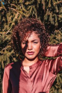 black woman with sun shining on her face, arm behind her head, wearing sateen deep pink blouse , shoulder length wavy natural hair, standing against a natural green leafy background